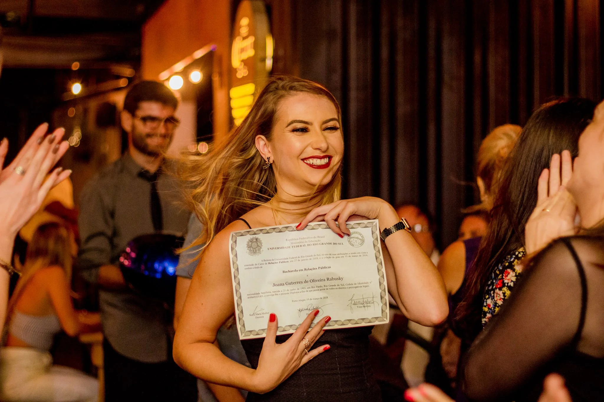 Mulher sorrindo segurando diploma de graduação em Relações Públicas, comemorando na cerimônia de formatura.