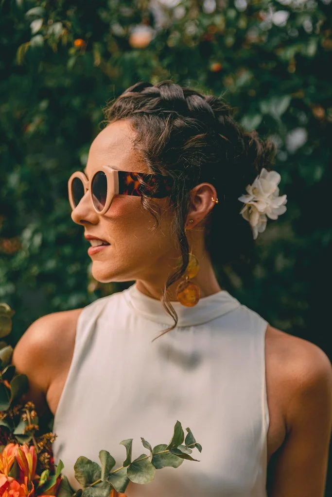 Mulher com cabelo preso, usando óculos de sol com armação tartaruga, vestido branco sem mangas, brincos de resina dourados, com flor branca no cabelo, rodeada de folhagens e flores, em ambiente ao ar livre.