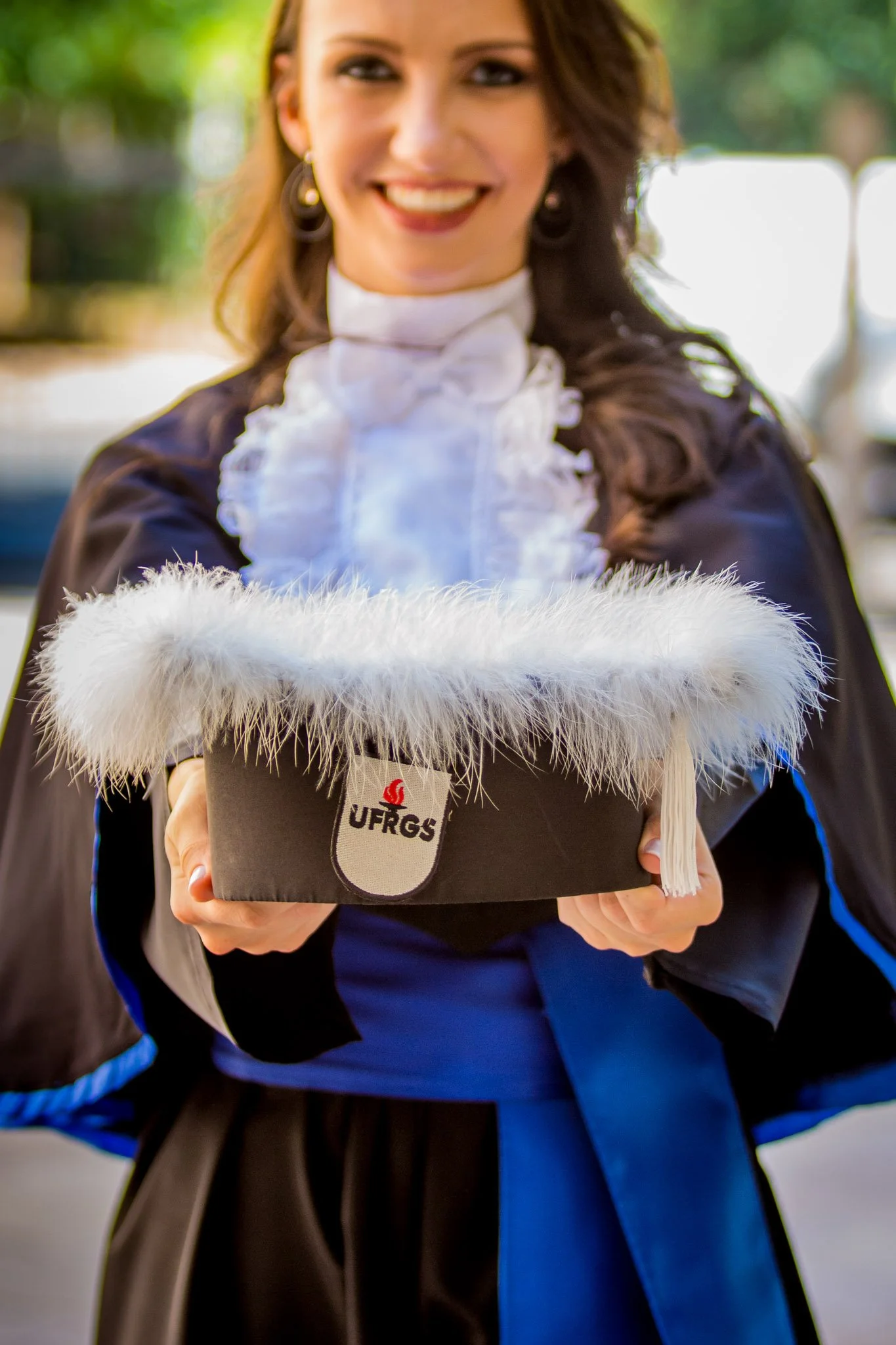 Mulher sorridente com roupa acadêmica de formatura, segurando um chapéu de formatura com detalhes em pele, com o logo UFRGS, ao ar livre.