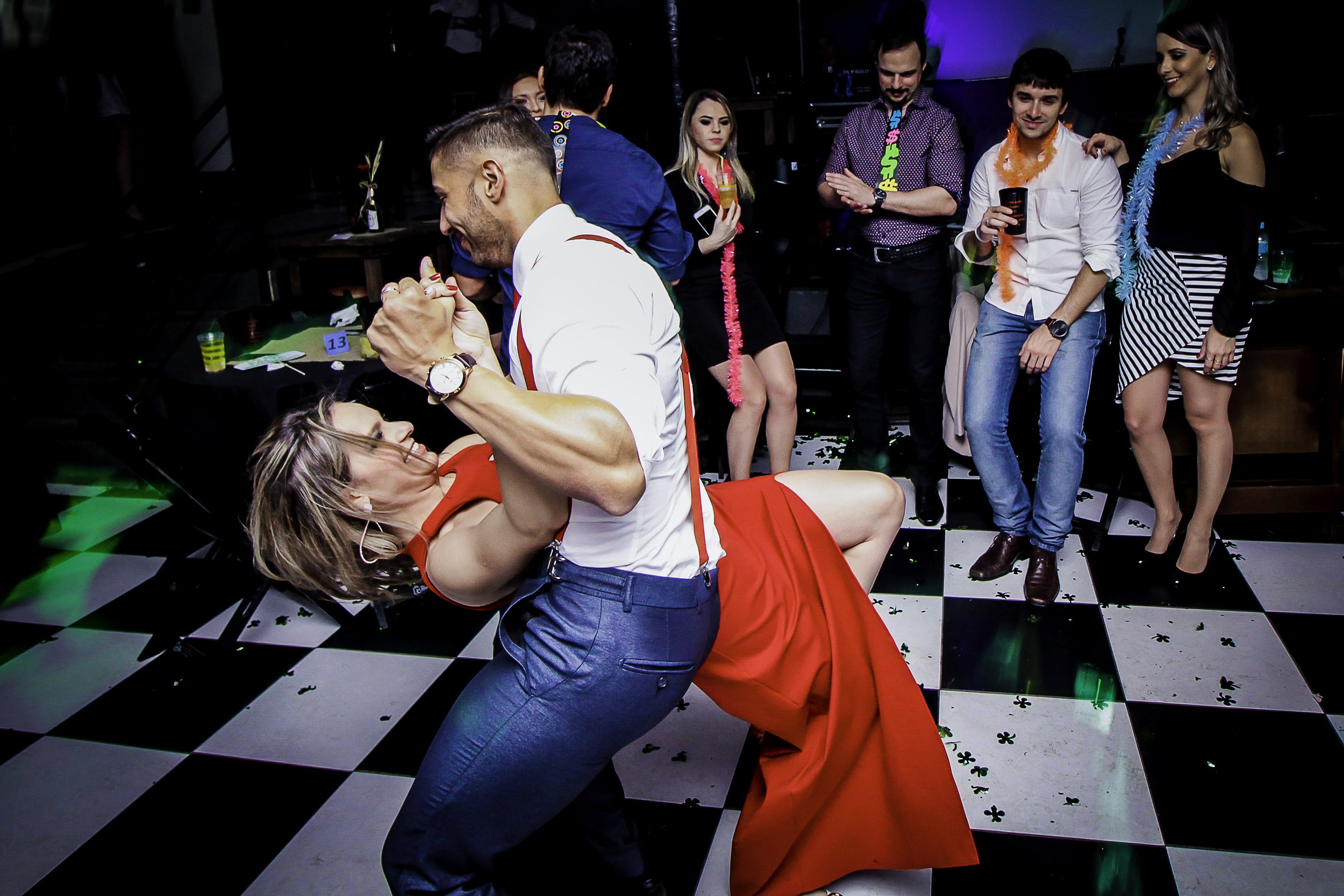 Pessoas dançando e se divertindo em uma festa, com um casal no centro fazendo uma dança de dança de salão, enquanto outros observam e se divertem ao redor.