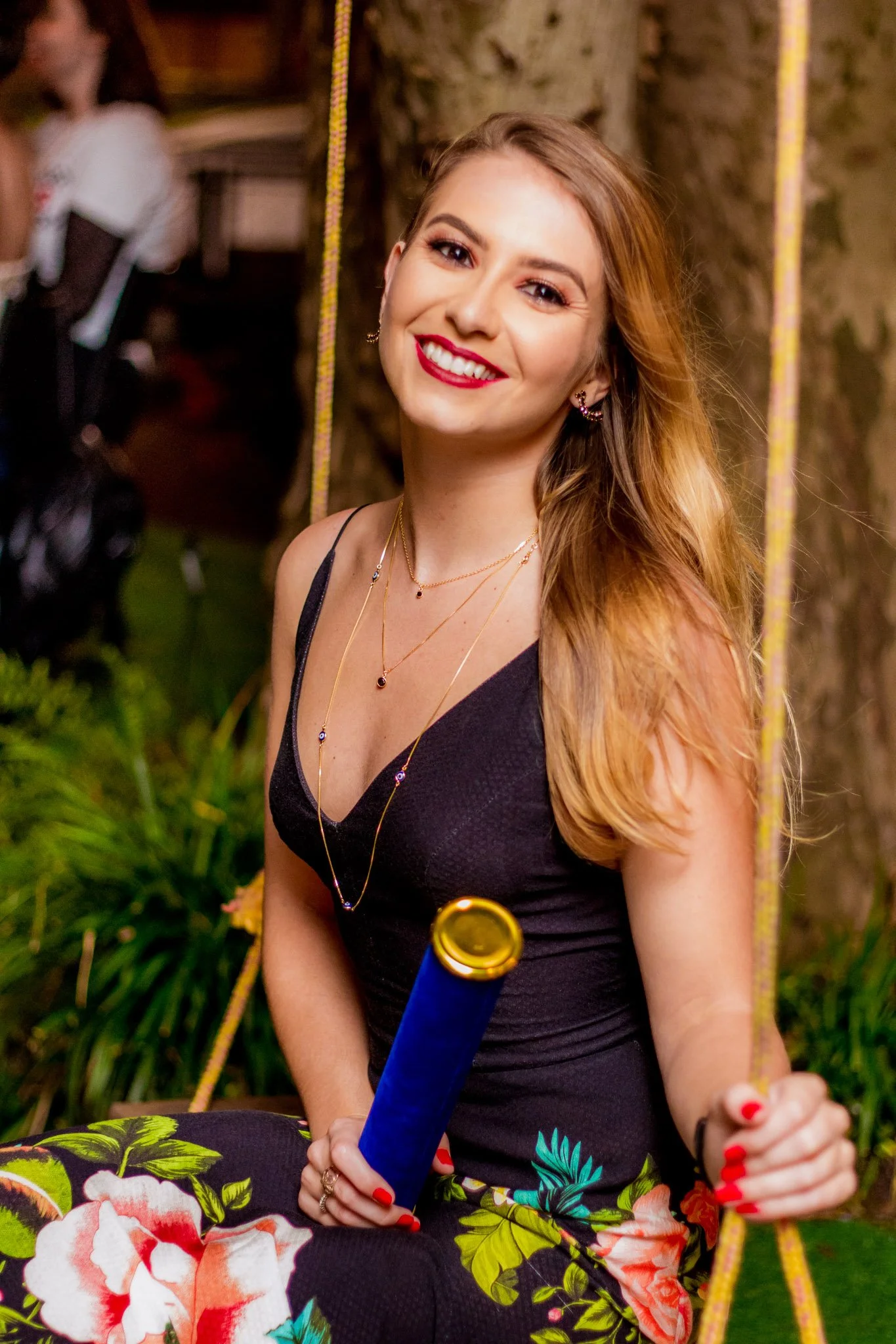 Jovem mulher sorridente sentada em uma gangorra de jardim, segurando uma rola de papel descartável azul e dourada, com cabelos castanhos, usando vestido preto e saia floral, com joias douradas e maquiagem vibrante em um ambiente ao ar livre.
