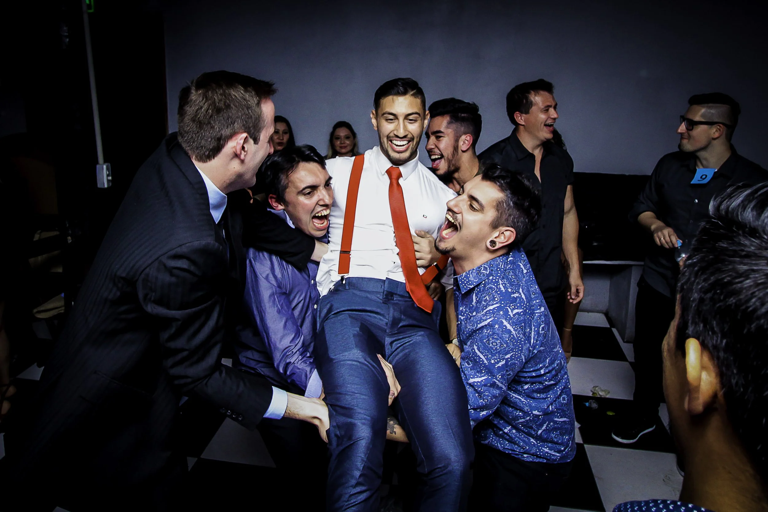 Grupo de pessoas sorrindo e rindo em torno de um homem que está sendo levantado ou apoiado por eles, numa festa ou celebração.