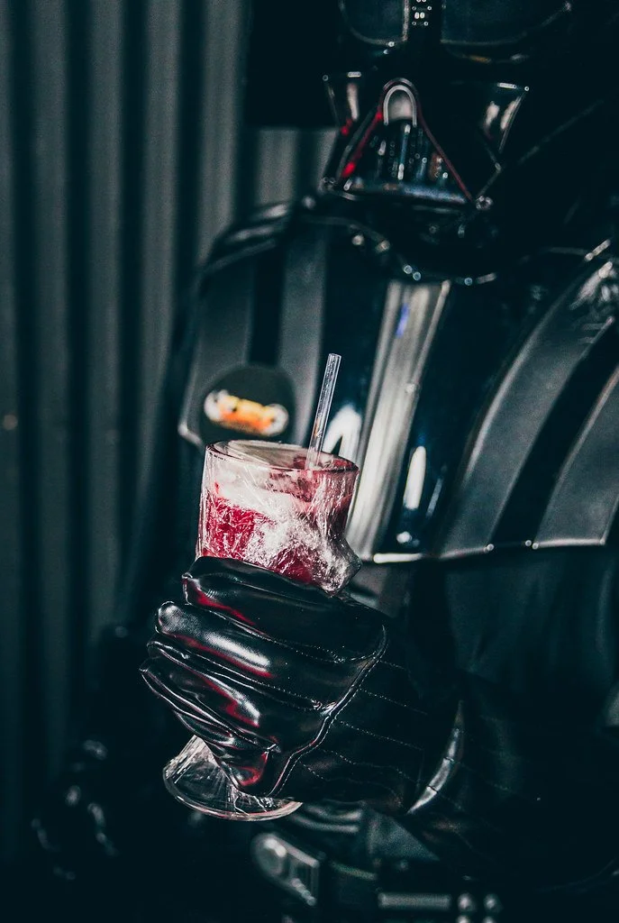 Mão com luva preta segurando um copo de vidro com drink vermelho, com gelo e canudo, ao lado de uma máquina de café espresso.