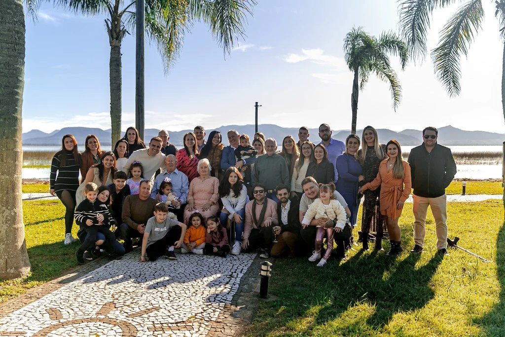 Grupo de pessoas posando ao ar livre sob árvores com cenário de montanhas ao fundo em um dia ensolarado.