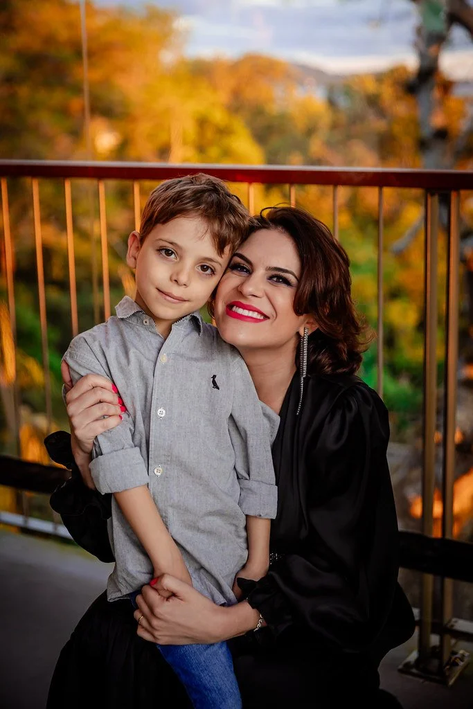Mulher sorridente segurando menino em um ambiente ao ar livre com árvores de folhas laranjas ao fundo.