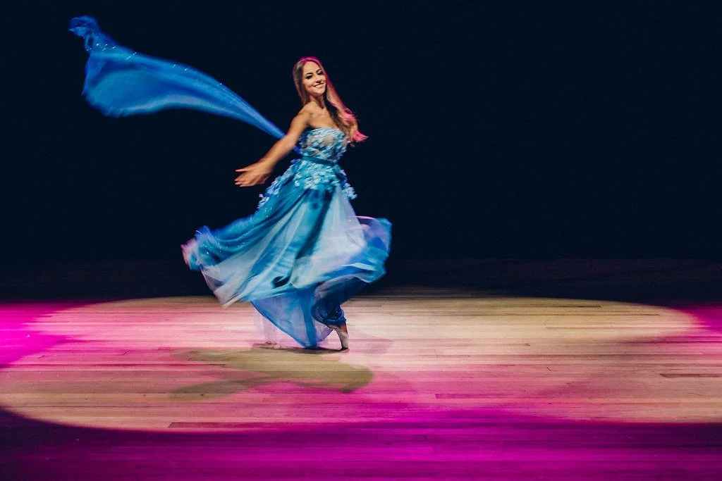 Mulher dançando em vestido azul em palco com iluminação rosa e fundo preto.