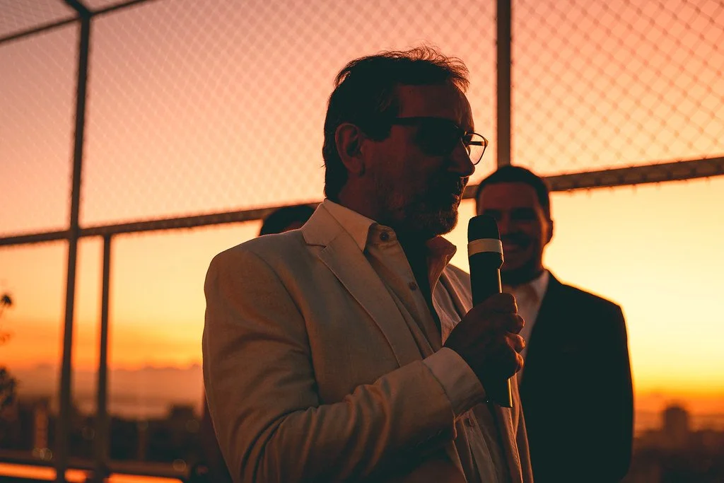 Homem com óculos escuros e cabelo grisalho fala ao microfone ao pôr do sol em uma área com mureta de metal.