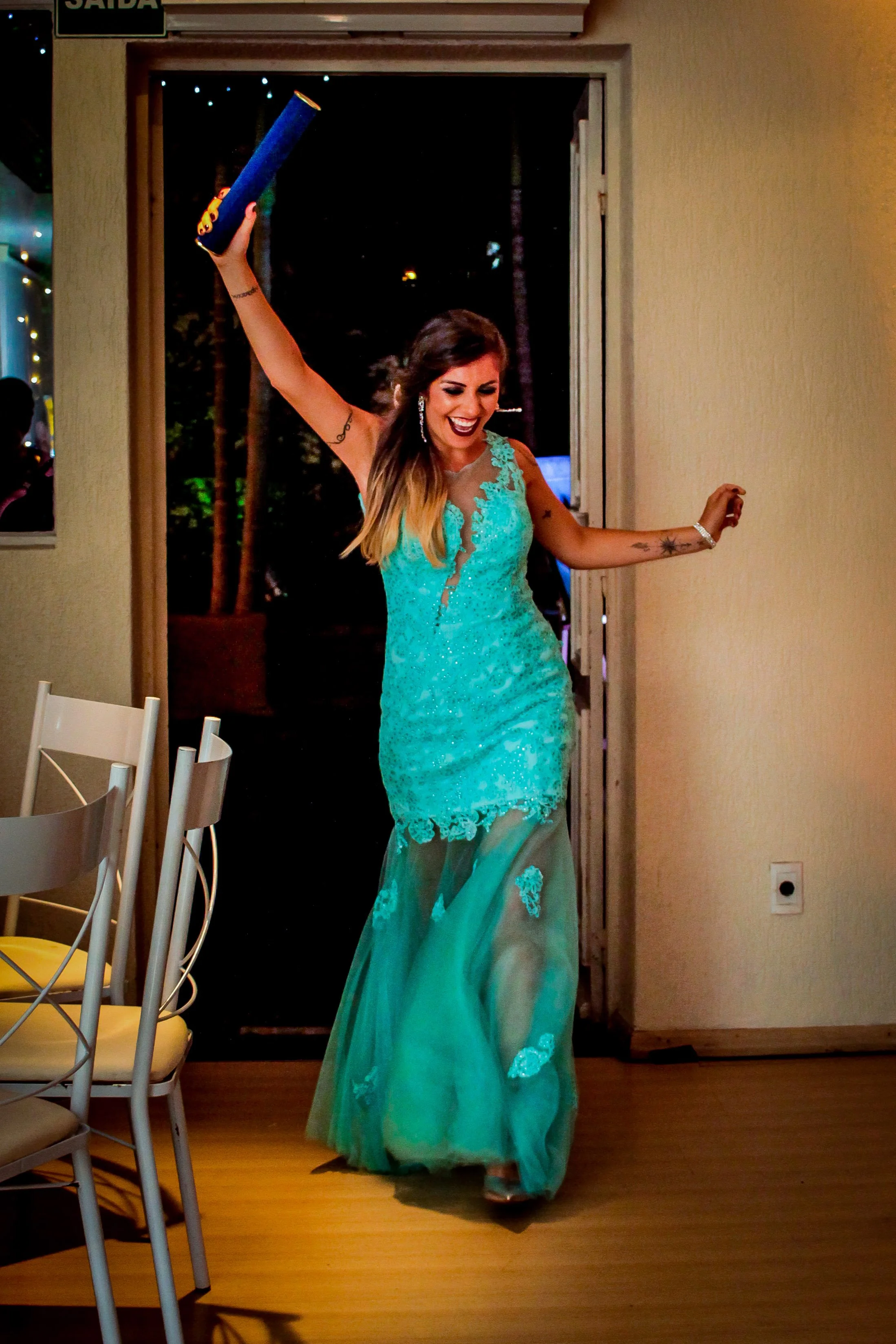 Mulher sorridente usando vestido longo azul, entrando com entusiasmo em um ambiente interno, possivelmente uma festa ou celebração, segurando um objeto azul na mão levantada.
