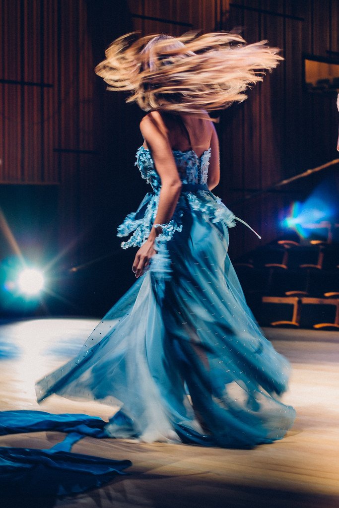 Mulher dançando em um palco com vestido azul longo e cabelo solto, em movimento.