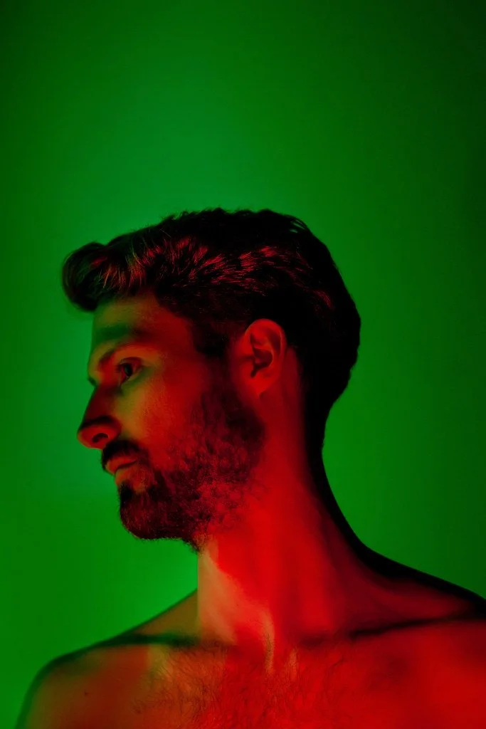 Retrato de um homem com barba, visto de perfil, com iluminação vermelha e fundo verde.