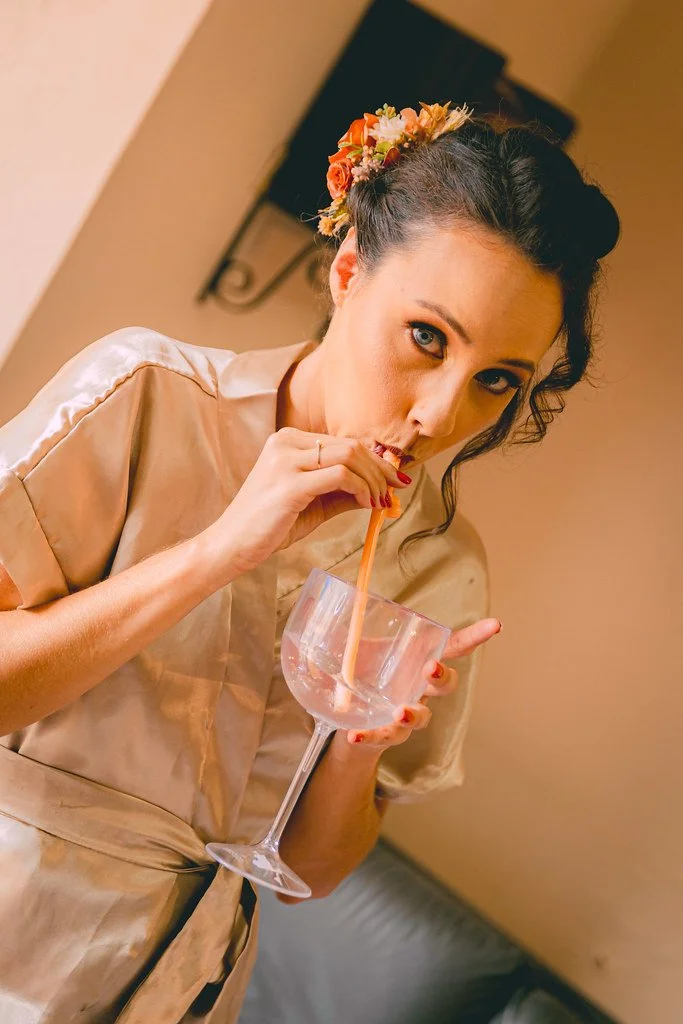 Mulher com cabelo escuro preso, usando vestido de seda bege, com arranjo de flores na cabeça, tomando uma bebida em um copo grande com canudo, em ambiente interno.
