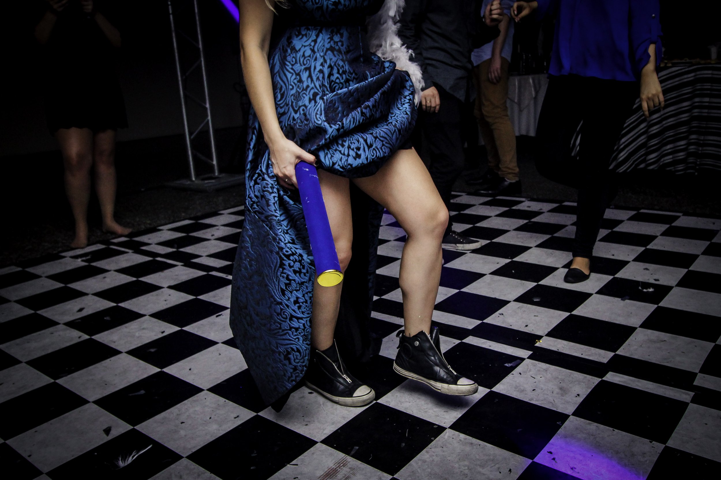 Pessoa com saia longa azul e tênis pretos, segurando um tubo azul, dançando em uma pista de dança estilo xadrez preto e branco, com outras pessoas ao fundo.