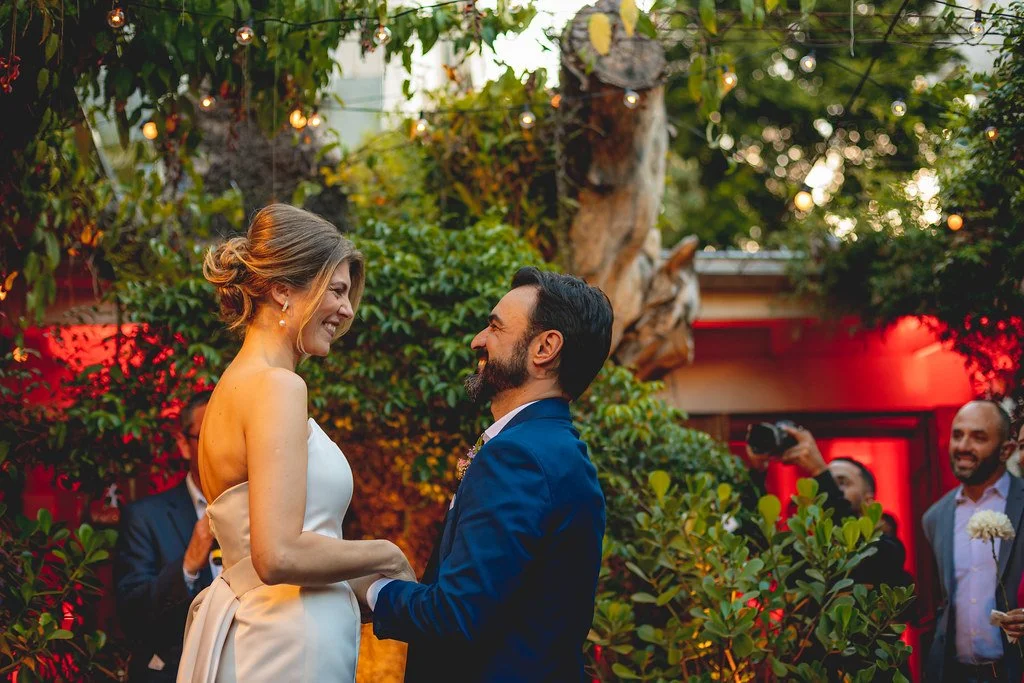 Casal em uma cerimônia de casamento ao ar livre, sorrindo e segurando as mãos um do outro, com convidados ao fundo e vegetação ao redor.