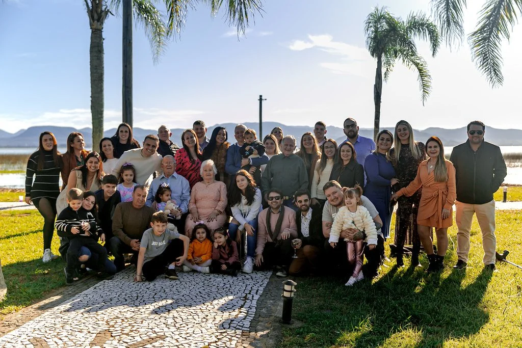 Grupo de pessoas posando ao ar livre próximo a uma praia e árvores, com montanhas ao fundo.