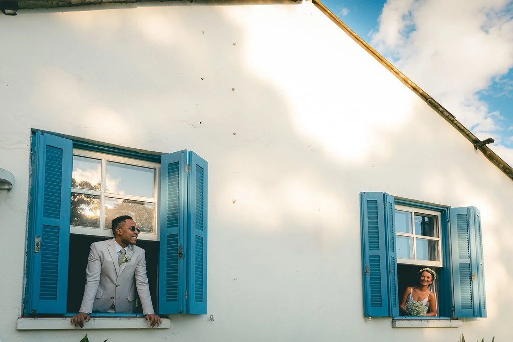 Casal sorridente com roupas de casamento, cada um na janela de uma casa com janelas azuis abertas.