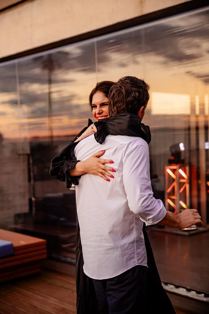 Homem e mulher se abraçando em ambiente moderno ao pôr do sol, com reflexo de céu laranja na parede de vidro.
