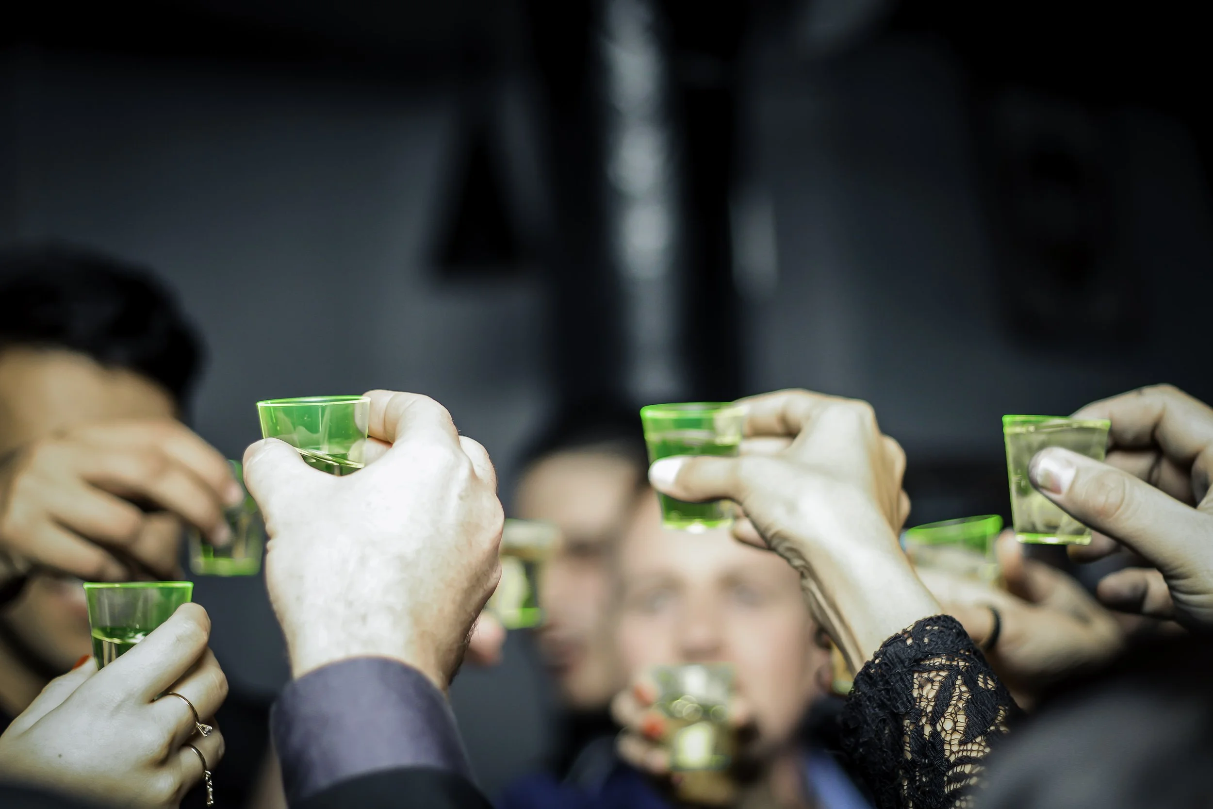 Pessoas brindando com pequenos copos verdes em uma celebração, com uma mulher ao centro com cabelo loiro ao fundo.