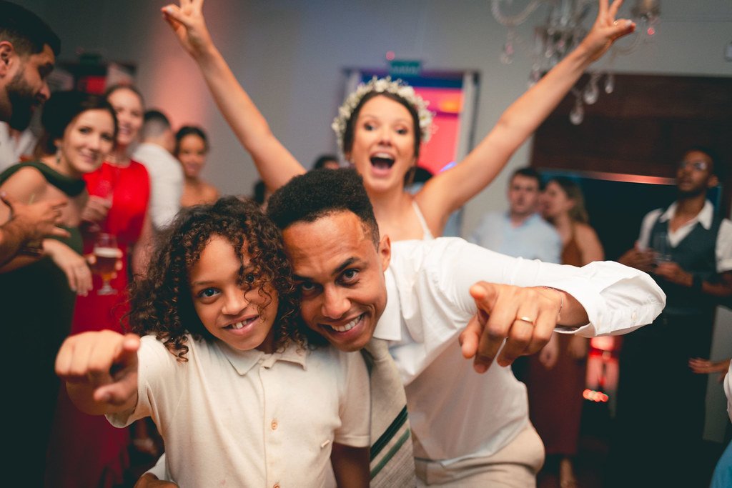 Pessoas comemorando em festa, com uma mulher usando uma coroa de flores e várias outras sorrindo e se divertindo ao fundo.