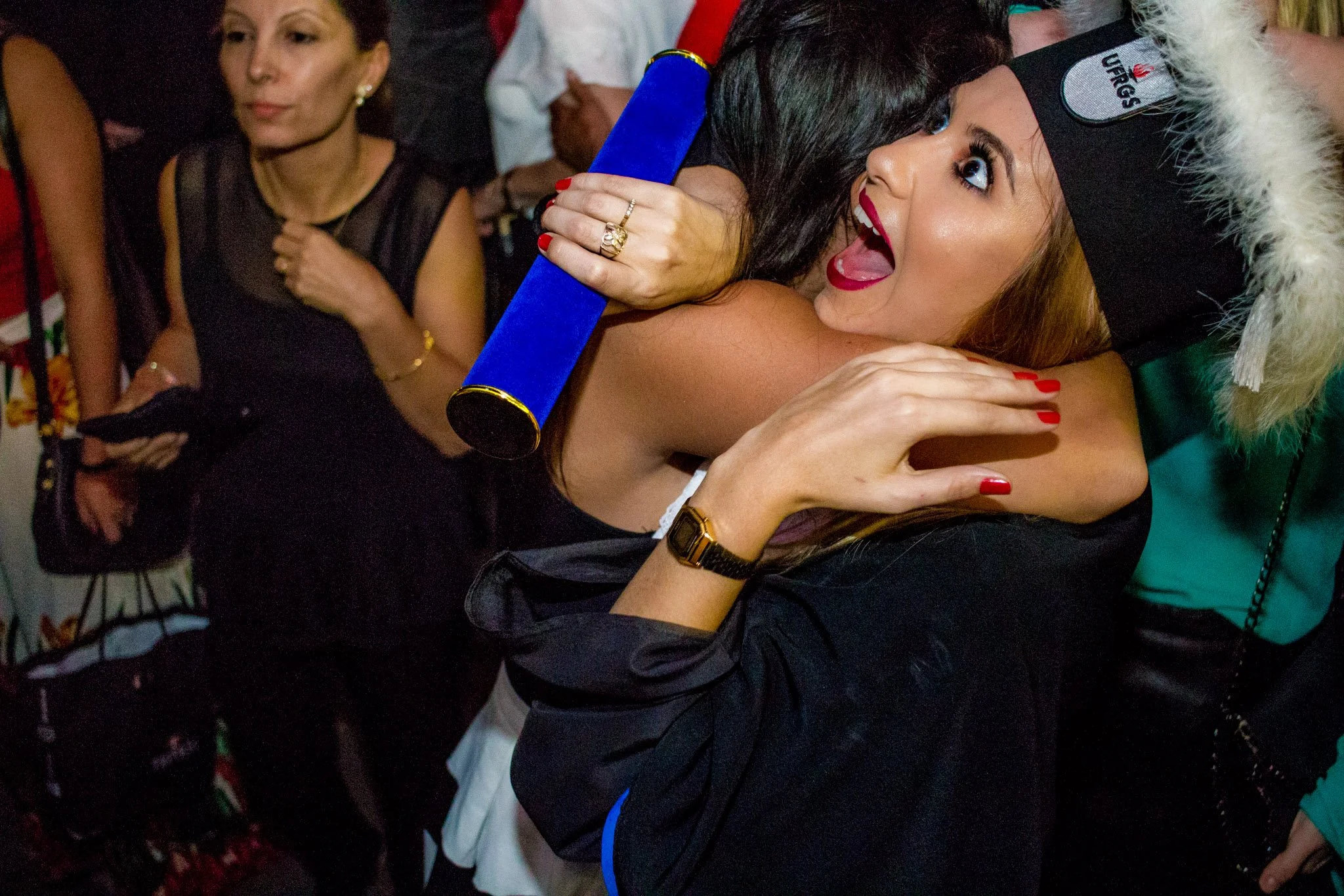 Mulher com chapéu de formatura e capa comemorativa, sorrindo e recebendo um abraço em uma celebração