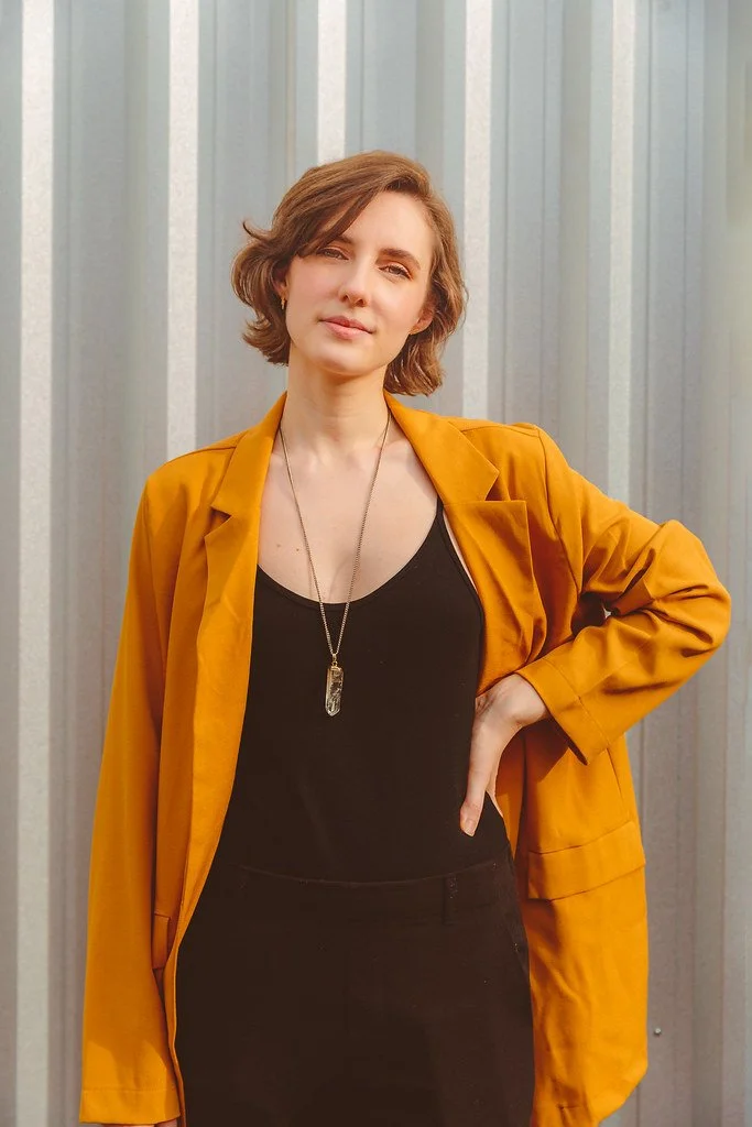 Mulher de cabelos curtos e castanhos, vestindo blusa preta, blazer laranja, e colar com pingente, posando com uma mão na cintura diante de fundo de painéis de metal.