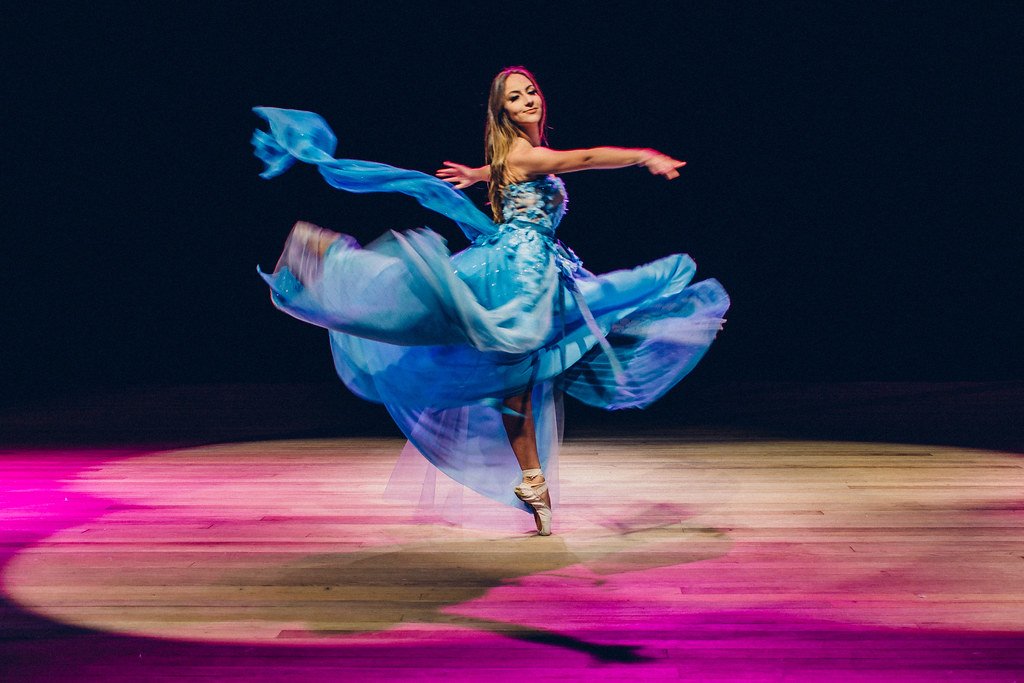 Bailarina vestida com vestido azul está dançando no palco, girando com movimento gracioso e sorrindo.