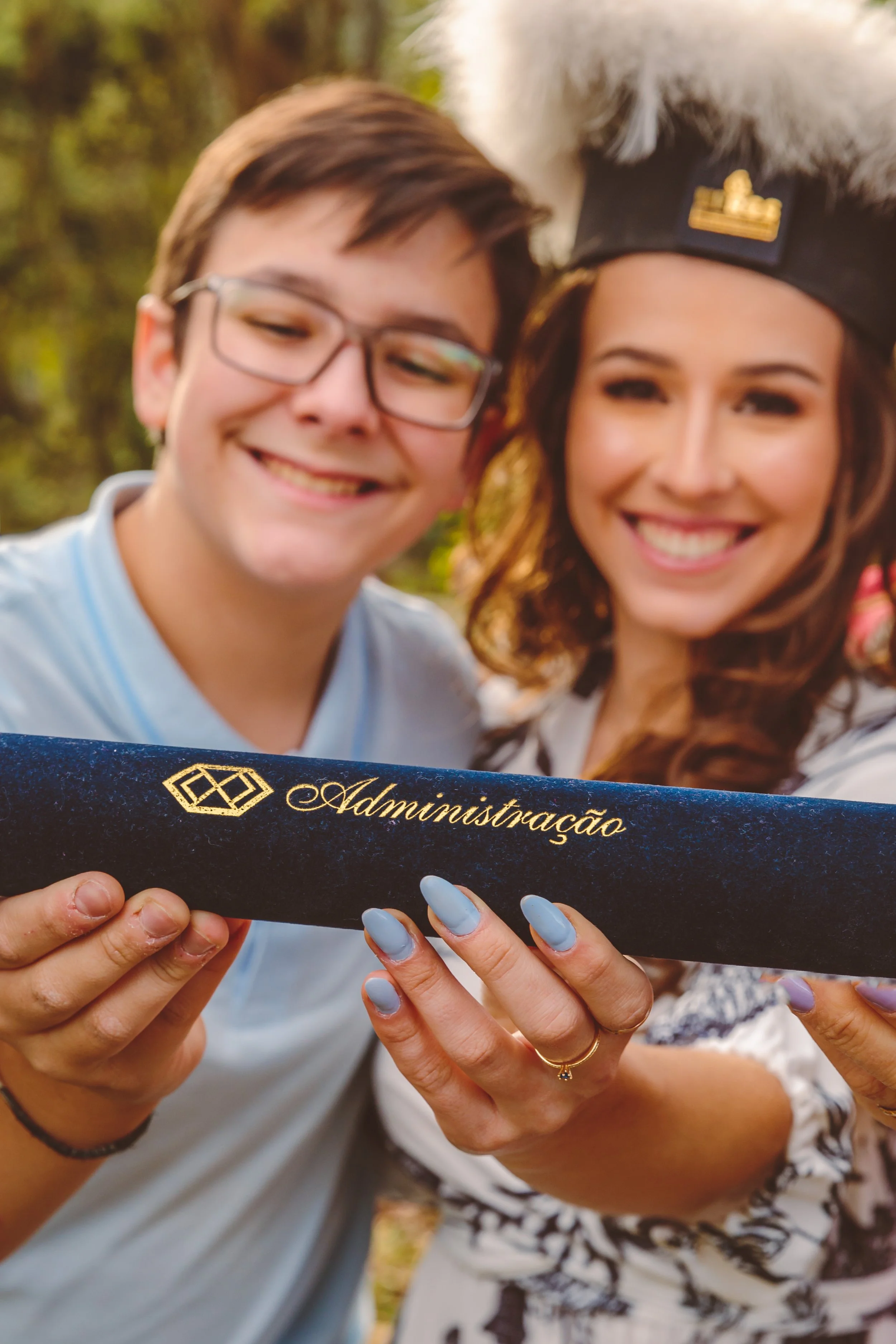 Retrato de uma mulher e um homem sorrindo segurando um diploma de graduação, que tem a palavra 'Administração' escrita, em um ambiente ao ar livre.