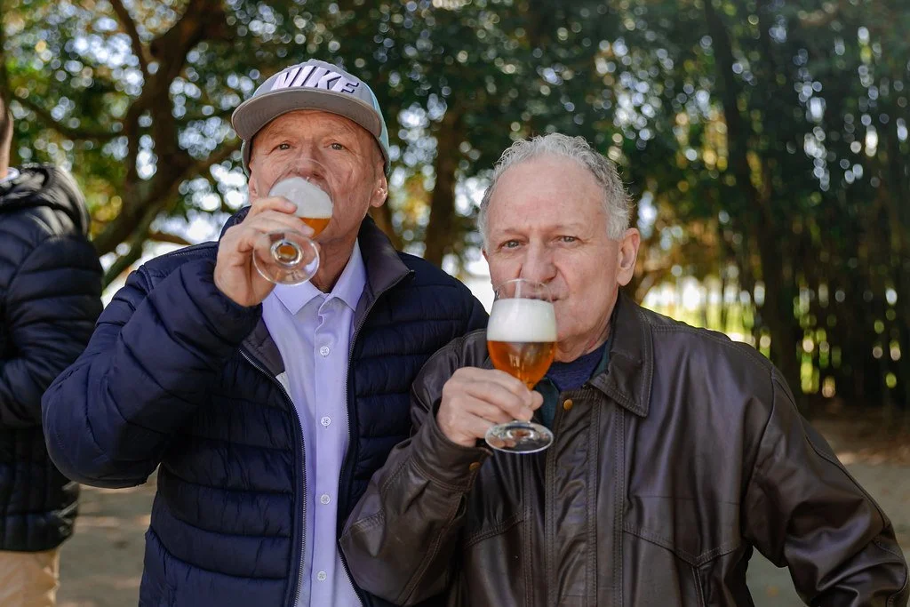 Dois homens idosos brindando com cervejas em um ambiente ao ar livre, com árvores ao fundo.