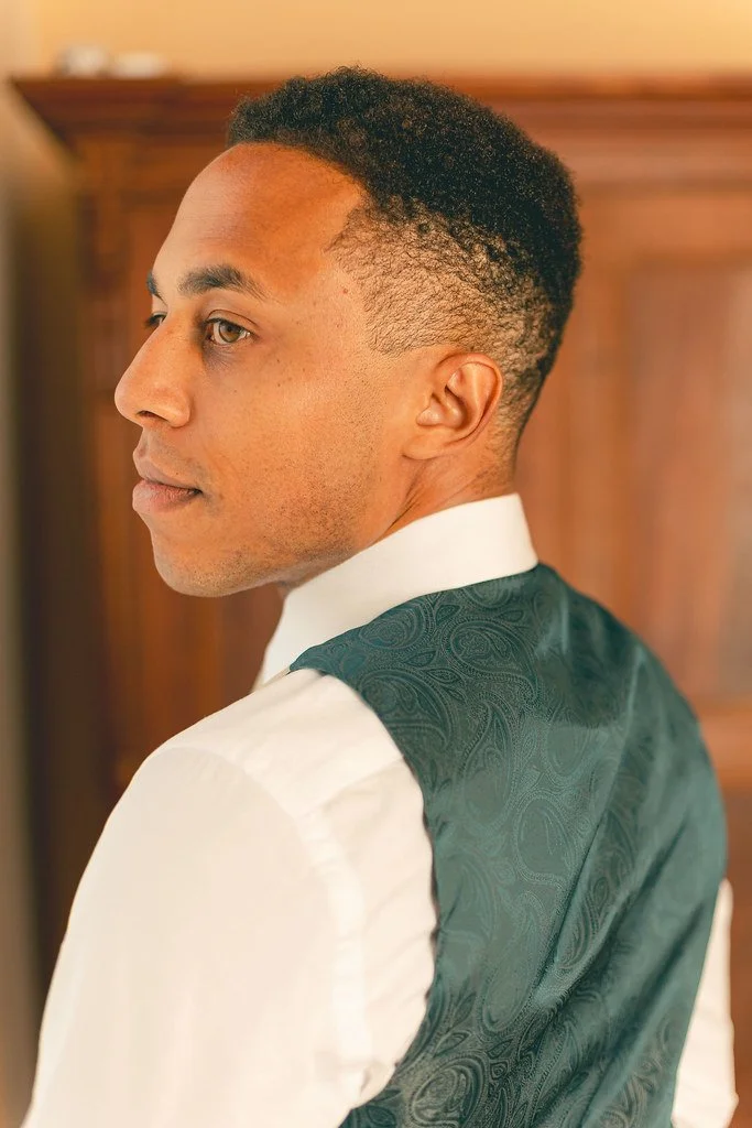 Homem com cabelo curto e liso, vestindo camisa branca e colete verde com estampa sutil, olhando para o lado em ambiente com móveis de madeira ao fundo.