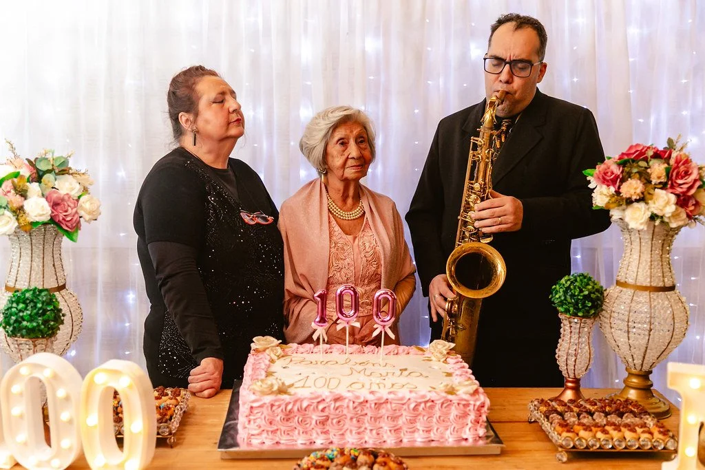 Três pessoas celebrando o 100º aniversário com um bolo rosa decorado, velas em forma de 100 e arranjos de flores, enquanto uma mulher toca saxofone.