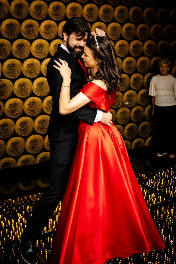 Casais dançando abraçados, com fundo decorativo de rolos de papel em tons de dourado, em ambiente elegante.