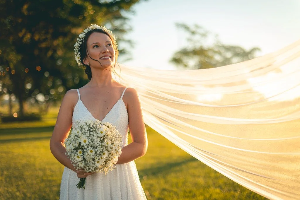 Noiva sorridente em um campo ao pôr do sol, usando vestido de casamento, com coroa de flores na cabeça, segurando buquê de flores brancas.