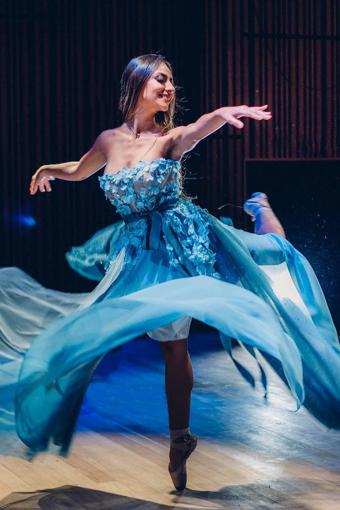 Bailarina em movimento com vestido azul em palco com fundo de painéis de madeira.