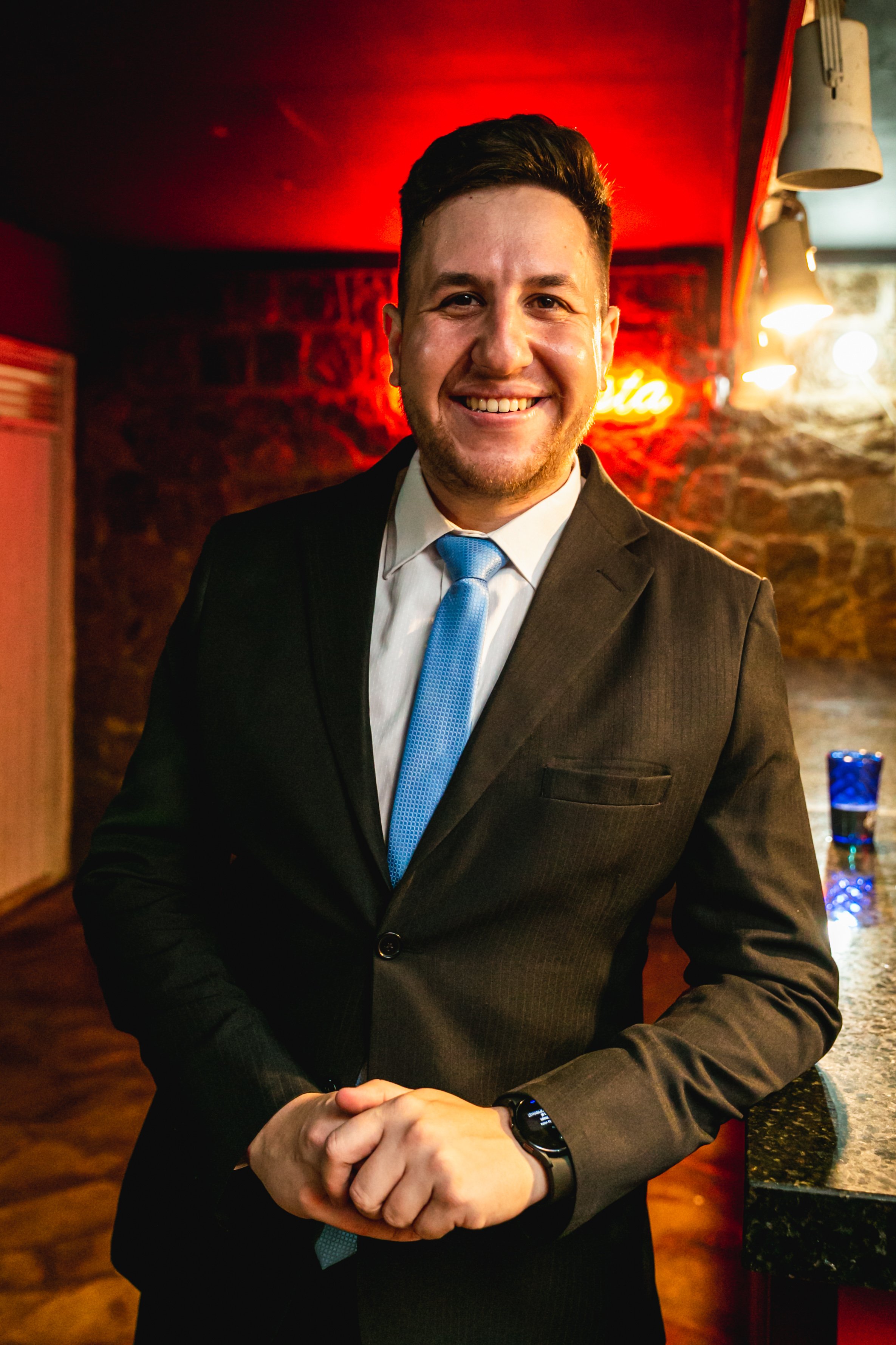 Homem sorridente com terno e gravata azul, de frente, em ambiente de bar ou restaurante com iluminação quente e parede de pedra ao fundo.