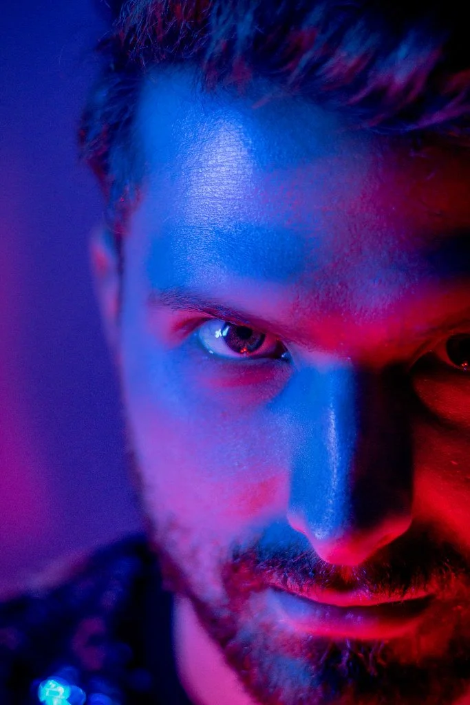 Close-up de um homem com iluminação azul e vermelha, focando no rosto e olhos.