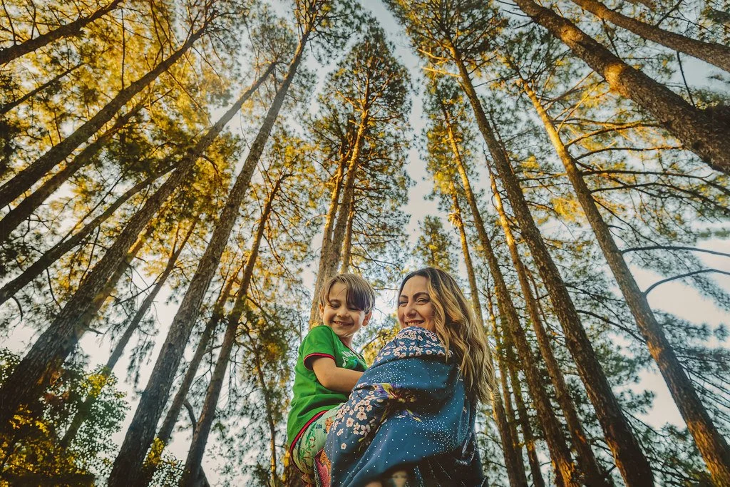 Uma mulher segurando uma criança em um bosque de árvores altas, com o céu ao fundo, durante o pôr do sol.