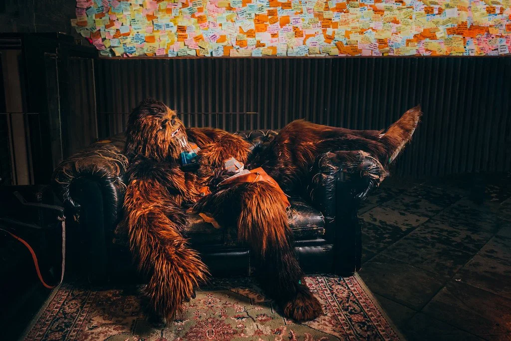 Pessoa de fantasia de cachorro de pelos longos e castanhos, deitada e com máscara de oxigênio, em um sofá de couro preto, em um ambiente com parede cheia de papéis adesivos coloridos.