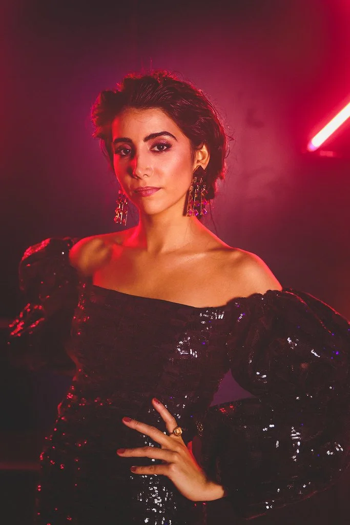 Mulher com cabelo escuro e maquiagem sofisticada, vestindo vestido preto com brilho e acessórios dourados, posando com uma mão na cintura em ambiente com iluminação vermelha e neon