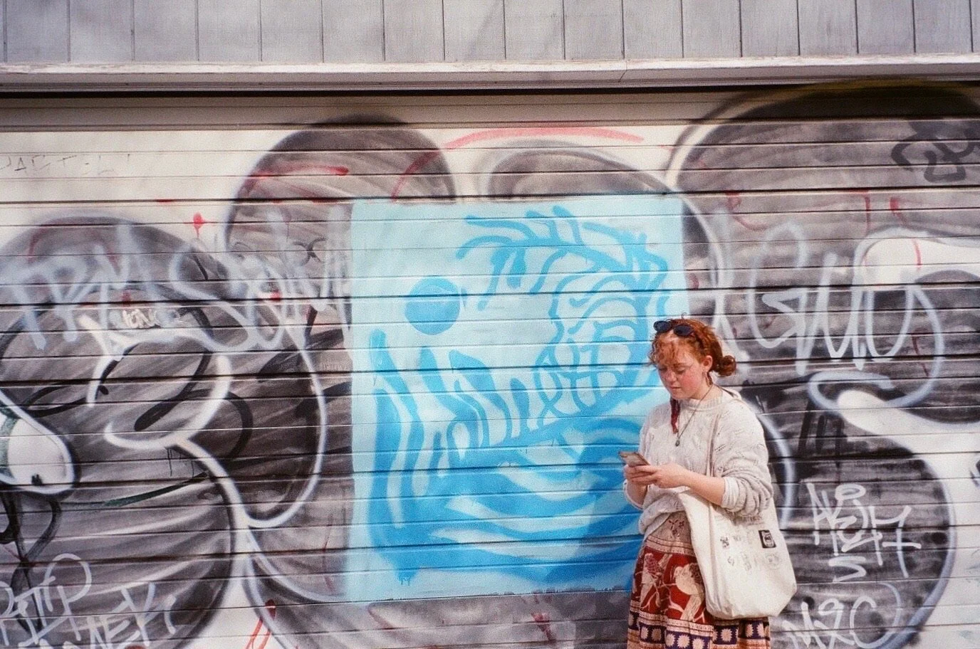 A woman with curly red hair, sunglasses on her head, wearing a beige sweater and patterned skirt, stands in front of a graffiti-covered garage door, looking at her phone.