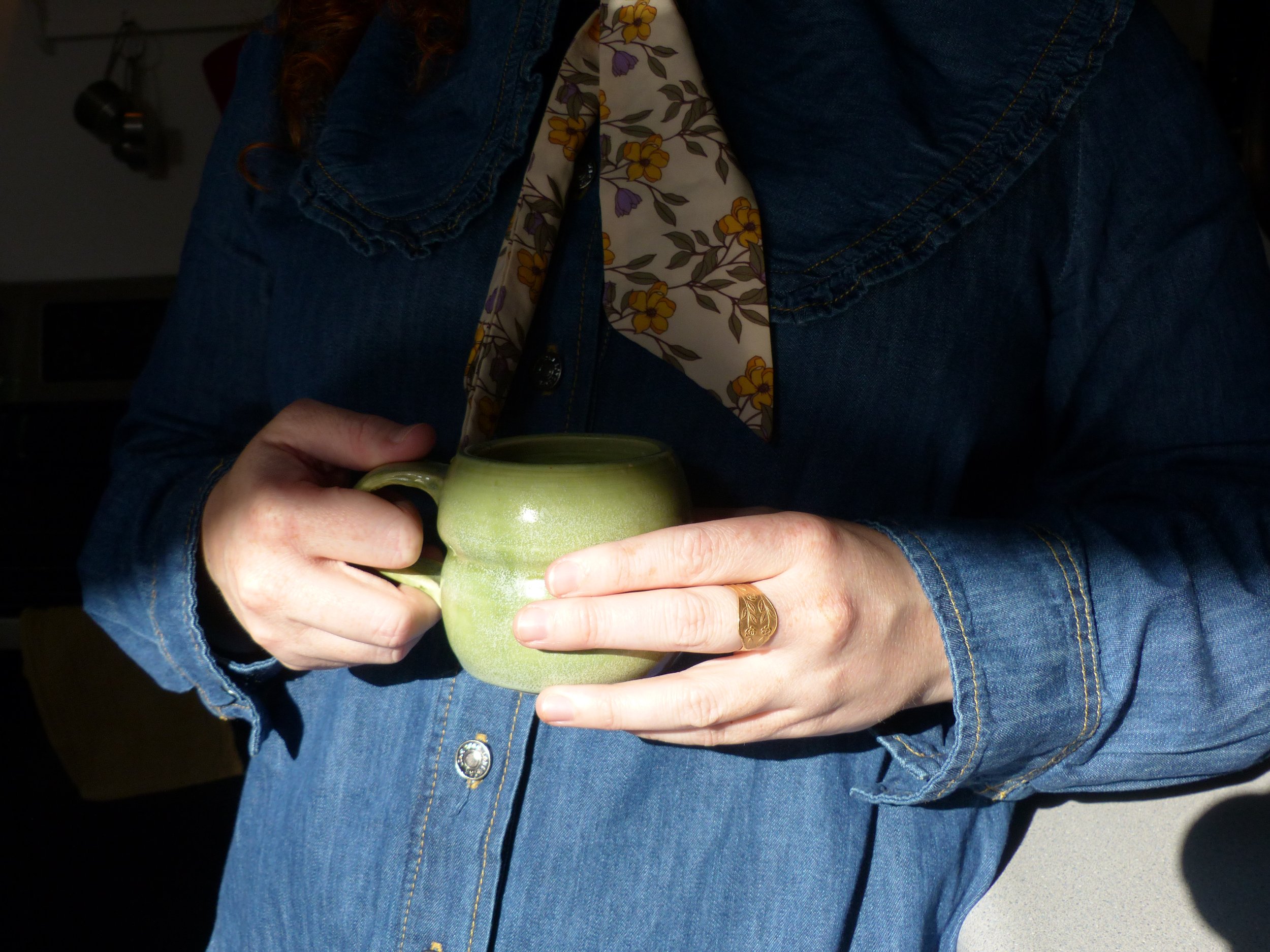 Person holding a green ceramic mug, wearing a denim shirt and a floral scarf.