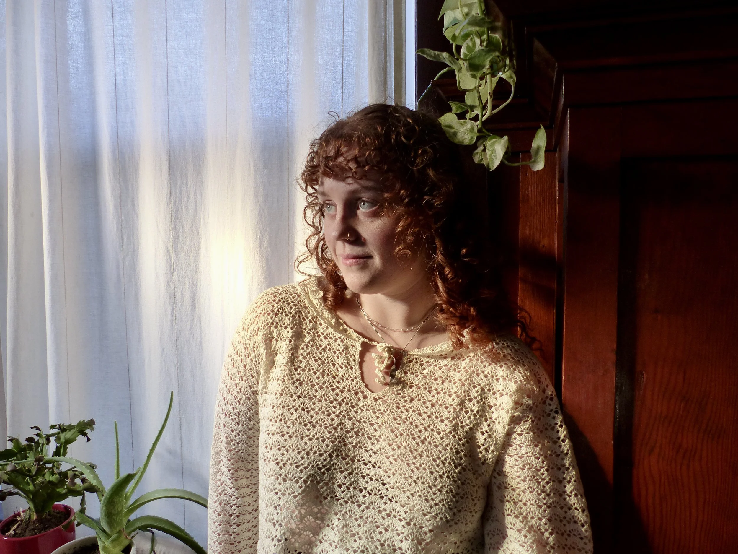 A woman with curly red hair standing by a window with sheer curtains, looking to the side, wearing a beige crochet top, with green houseplants nearby, and warm lighting.
