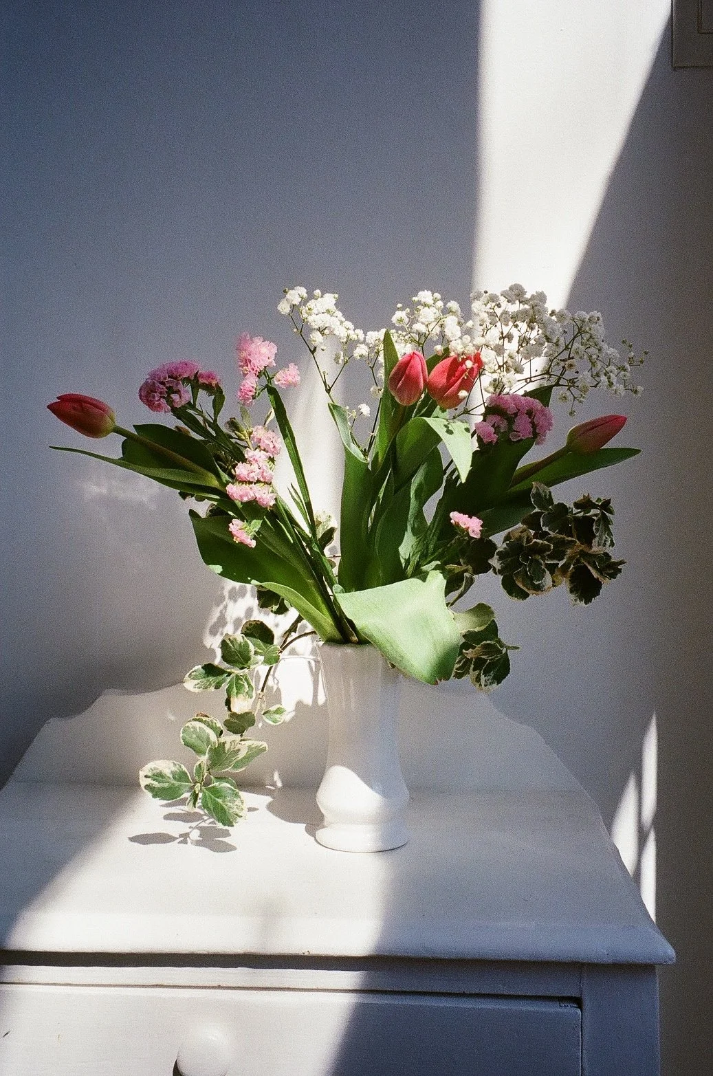 A white vase holding a mixed flower bouquet with pink tulips, pink and white carnations, white baby's breath, and variegated leaves, placed on a white surface with sunlight creating shadows.