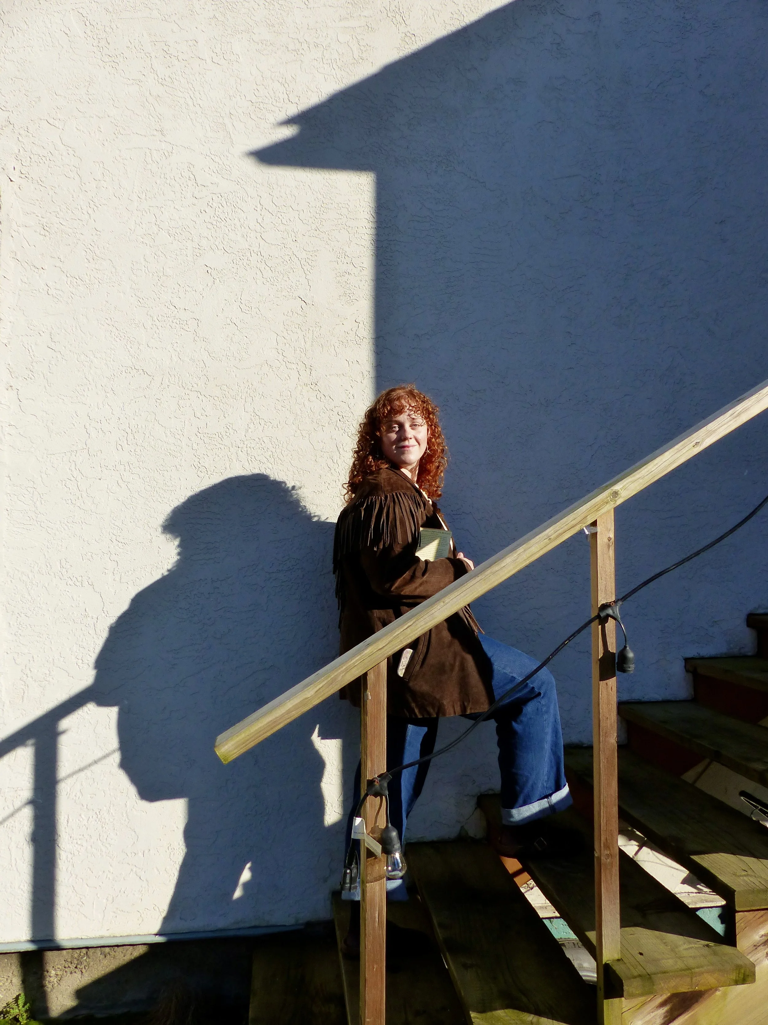 Woman with curly red hair sitting on stairs next to a light-colored textured wall, with her shadow cast on the wall, holding a book in her hands.