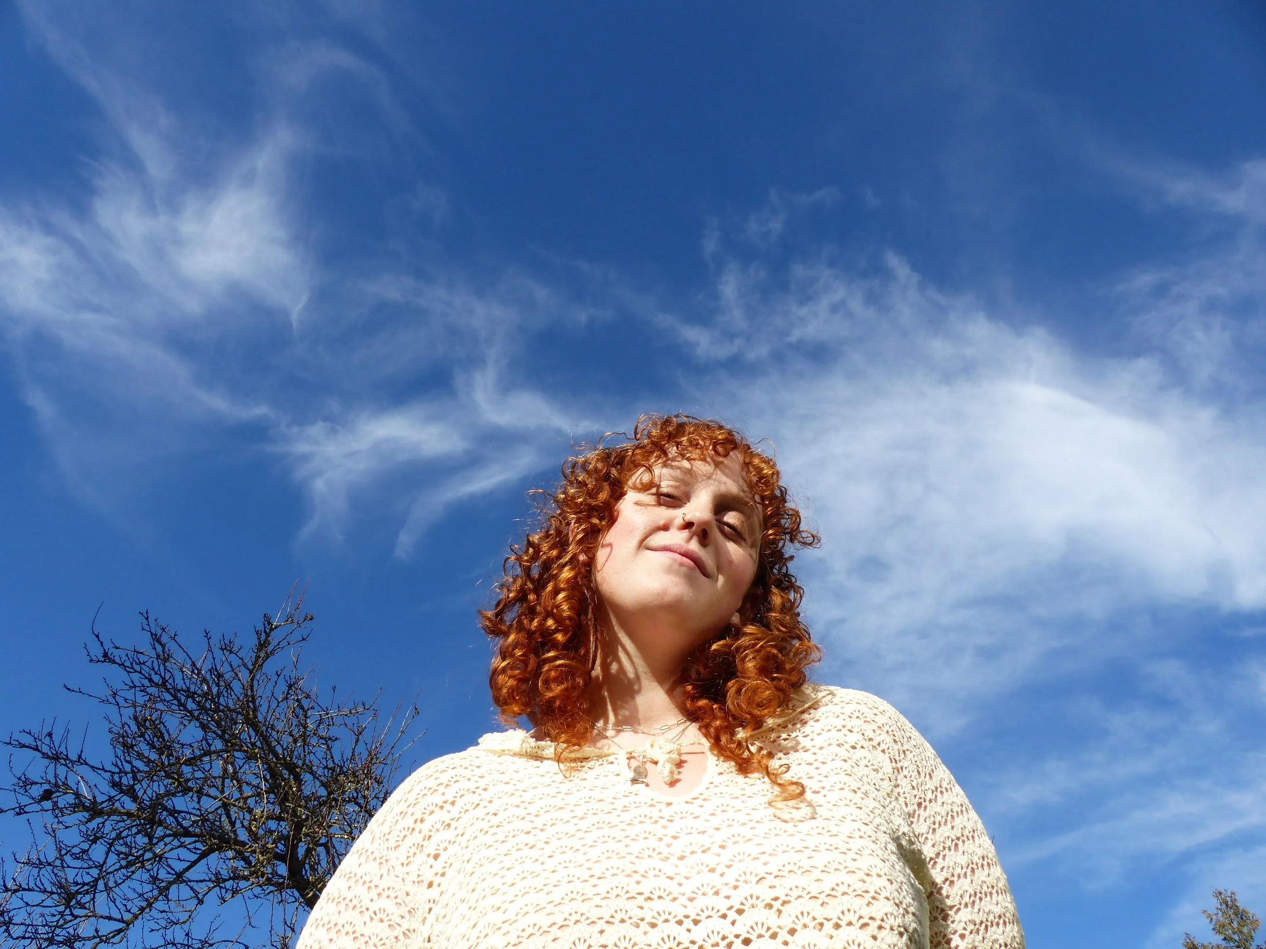 A woman with curly red hair standing outdoors against a bright blue sky with wispy white clouds, smiling confidently.