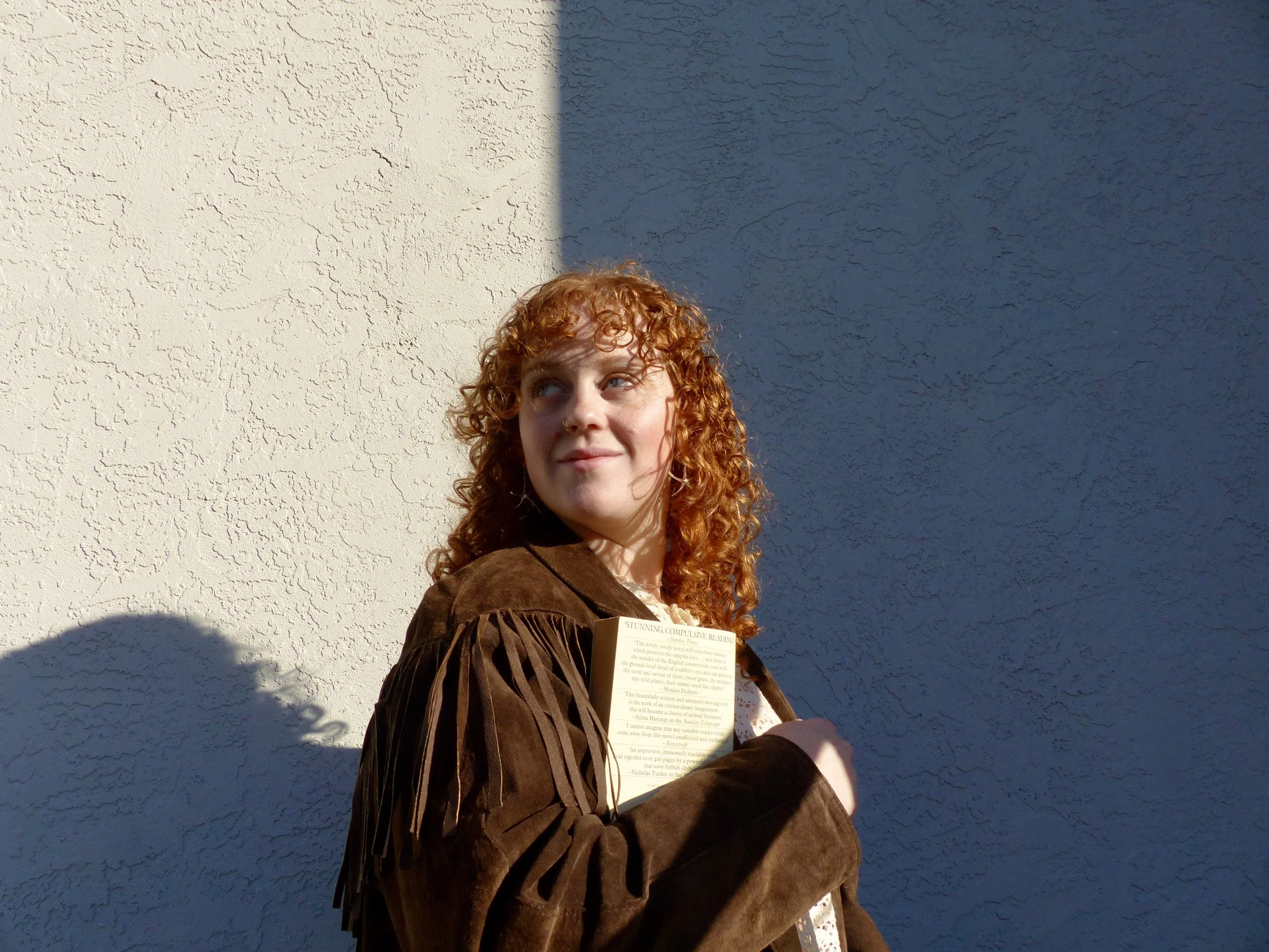 A young woman with curly red hair holding a book, standing against a textured wall with shadow cast on it, looking slightly upward and to the side.
