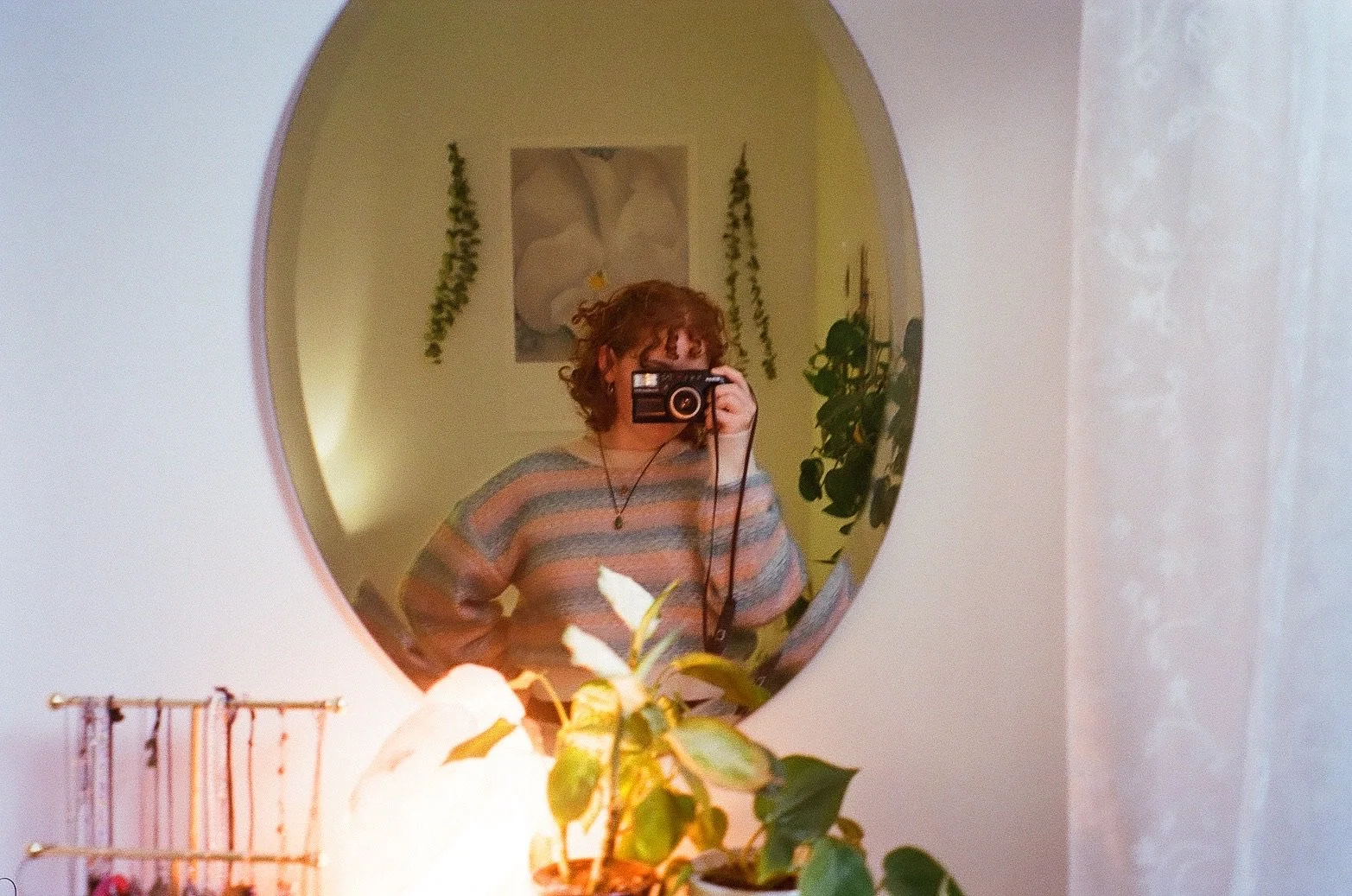 A woman with curly red hair taking a photo in a round mirror. She is wearing a striped sweater and a necklace. There are plants and a flower picture in the background.
