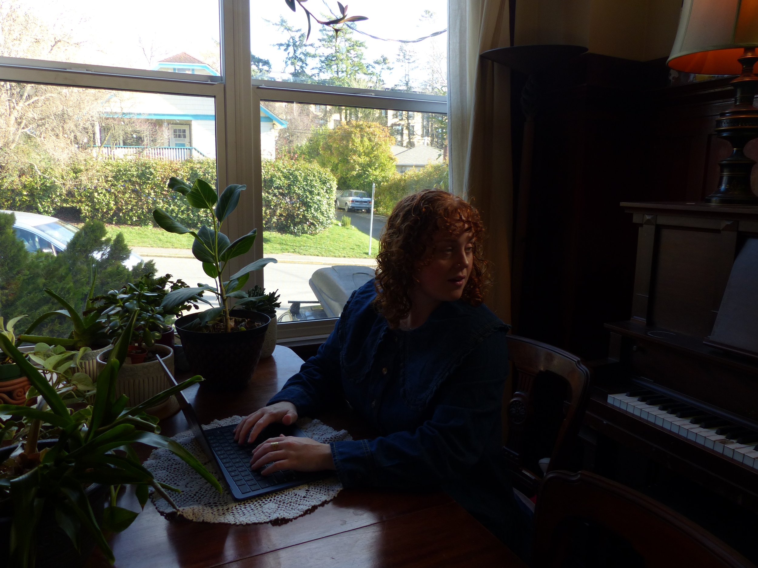 A woman with curly red hair sitting at a wooden table in a living room, using a keyboard computer. There are potted plants on the table, and a large window behind her reveals a suburban street with trees, houses, and parked cars.