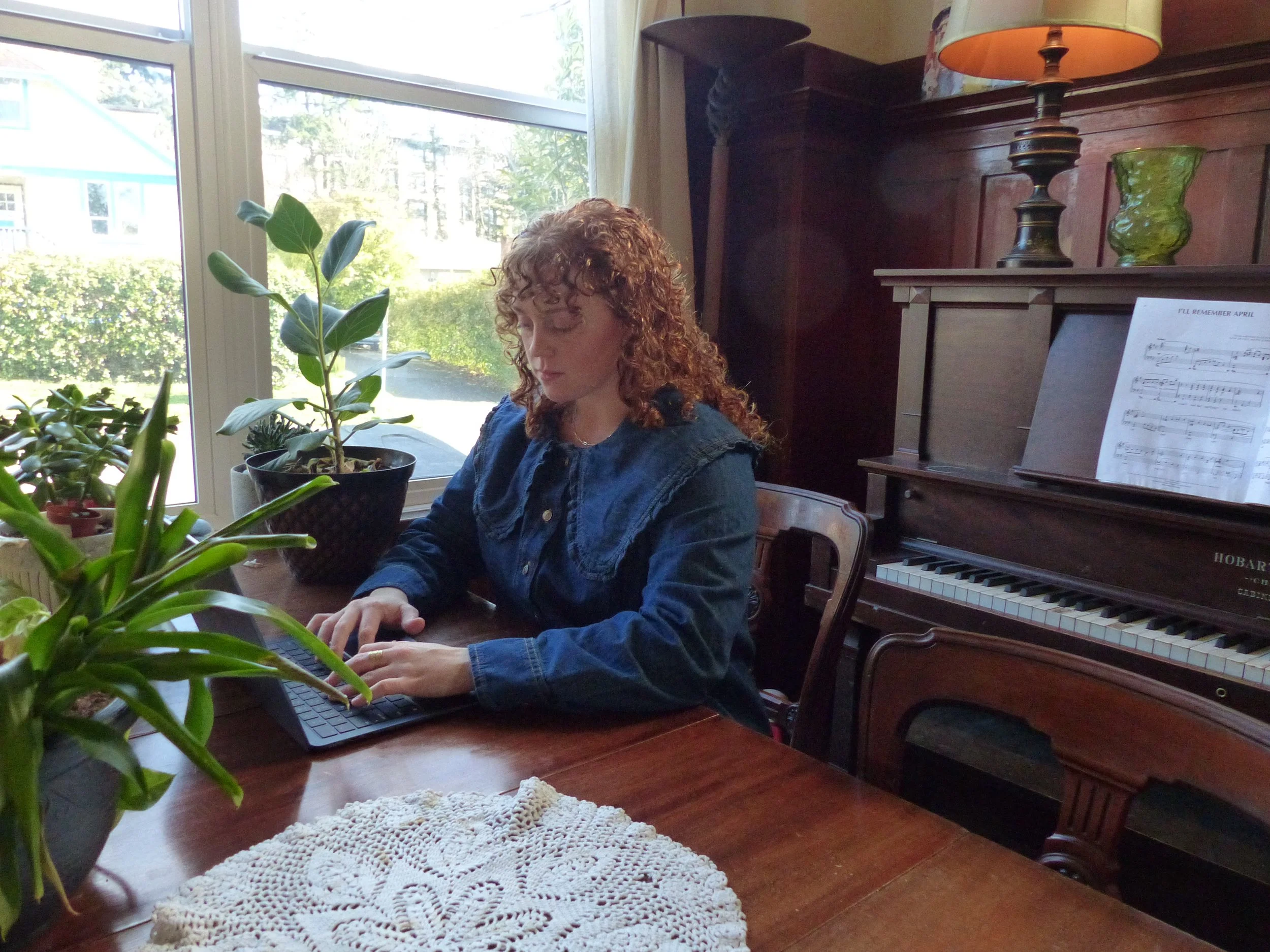 A woman with curly red hair wearing a denim shirt sits at a wooden table, working on a laptop, in a room with potted plants and a piano.