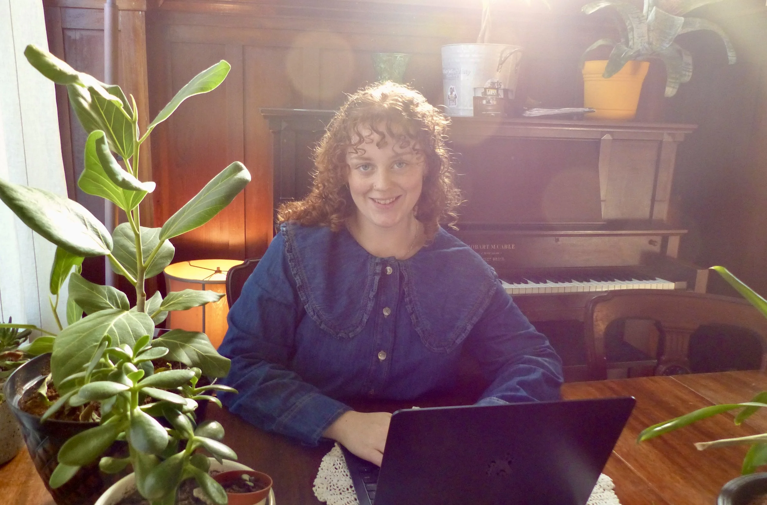 A woman with curly red hair sitting at a wooden table with a laptop, surrounded by green potted plants, in a room with wooden walls and a vintage piano in the background.