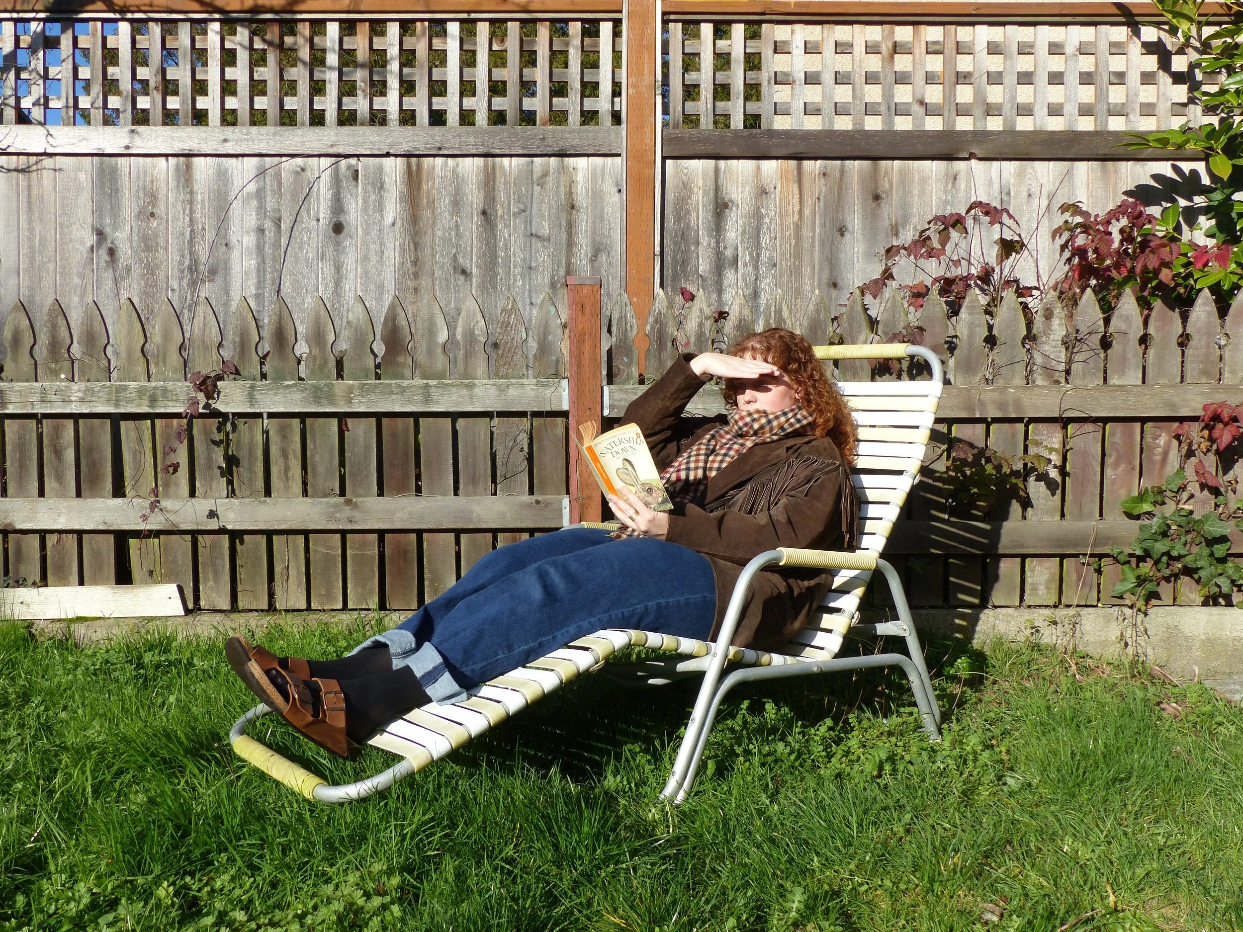 A woman with curly red hair wearing sunglasses, a brown jacket, and a plaid scarf, relaxing on a striped lounge chair in a backyard, reading a book while shielding her eyes from the sun with her hand.
