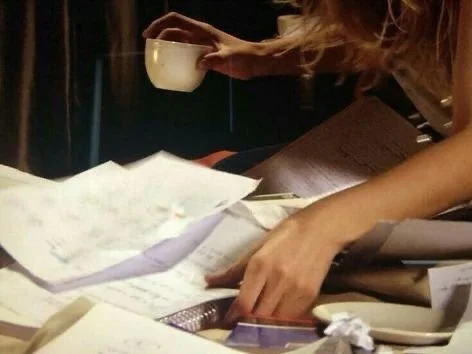 Person's hand reaching for a white cup on cluttered desk with papers and a notebook.